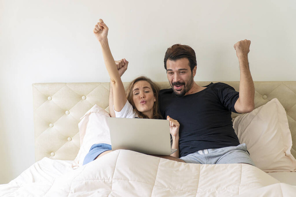 Életmód szórakoztató szabadidő koncepció. Izgatott fiatal pár viselni alkalmi ül együtt laptop, miközben éljenzés foci játék online vagy látszó hálózat jó hír az ágyban otthon.  - Fotó, kép