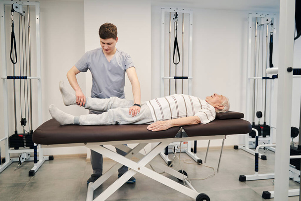 Physical therapist pulling old male right leg during treatment activity - Valokuva, kuva