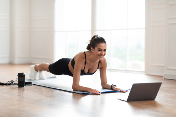 Junge fröhliche Frau beim Planken auf Matte mit Laptop - Foto, Bild