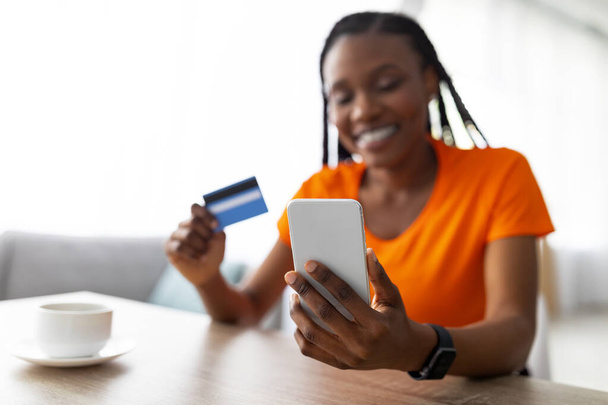 Aplikacja mobilna na zakupy. Czarna dama korzystająca ze smartfona i karty kredytowej, siedząca w kawiarni, kupująca towary w sklepie internetowym - Zdjęcie, obraz