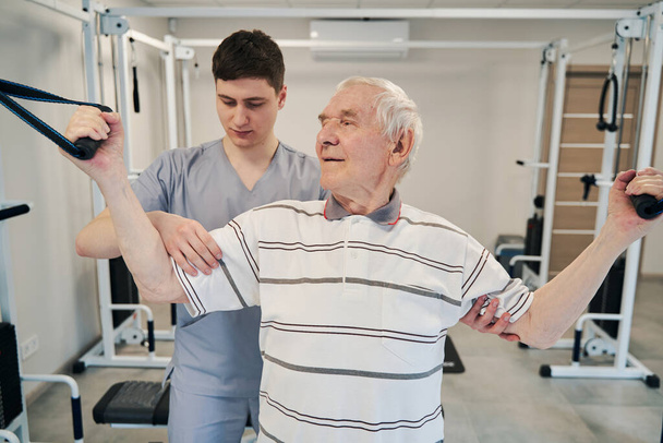 Rentner trainiert an Aufhängevorrichtung, während Arzt seine Muskeln überprüft - Foto, Bild