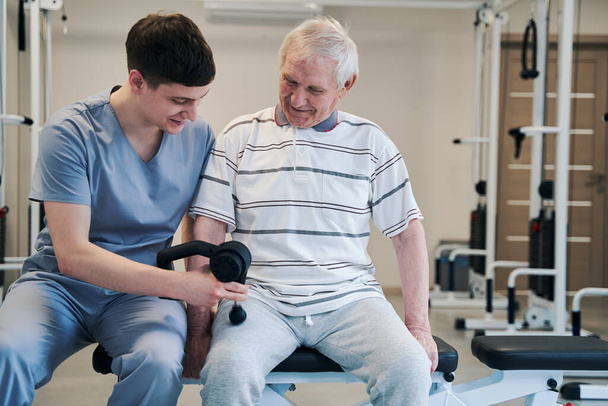 Fizjoterapeuta zapewniający leczenie nóg seniorowi z pistoletem do masażu - Zdjęcie, obraz