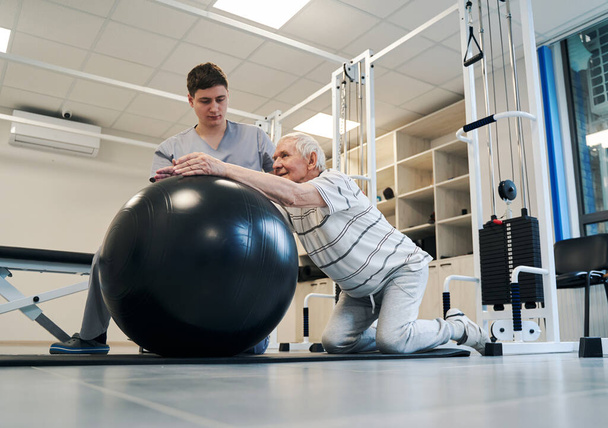 Pensioner standing on knees while putting arms on exercise ball - Fotó, kép