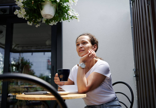 Gyönyörű vegyes verseny nő ül egy elegáns kávézó egy nyári teraszon, és úgy néz ki, figyelmesen a távolba egy csésze kávé a kezében - Fotó, kép