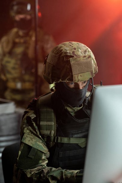 Muž v masce, helmě a maskovací uniformě při pohledu na monitor počítače - Fotografie, Obrázek