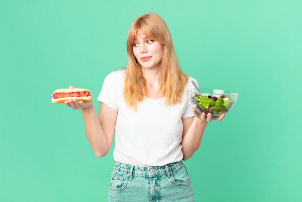 pretty red head woman holding a salad and a hot dog. diet concept - Foto, Imagem