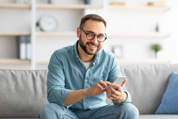 Fröhlicher Millennial-Typ in Brille sitzt auf gemütlicher Couch mit Handy und Nachrichten an seinen Freund, drinnen - Foto, Bild
