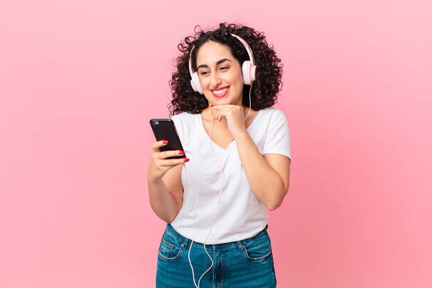 pretty arab woman smiling with a happy, confident expression with hand on chin with headphones and a smartphone - Foto, immagini