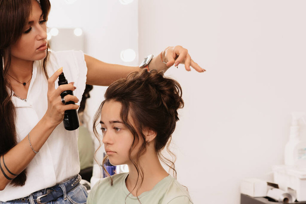 美容室での撮影。マスタースタイリストはスプレーヘアスプレーで固定の助けを借りて暗い髪の女の子の髪型を作ります - 写真・画像
