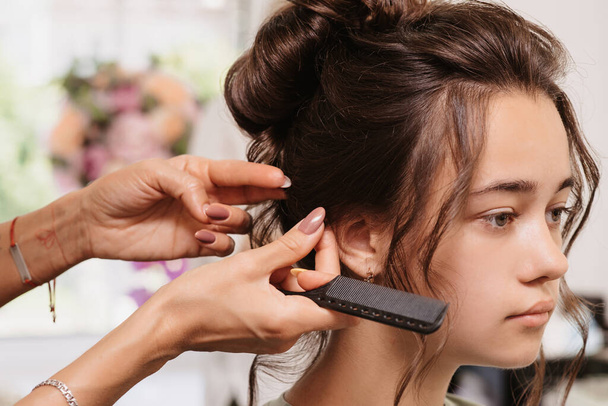 Tournage dans un salon de beauté. Un coiffeur corrige la coiffure d'une jeune fille aux cheveux bruns avec un peigne. - Photo, image