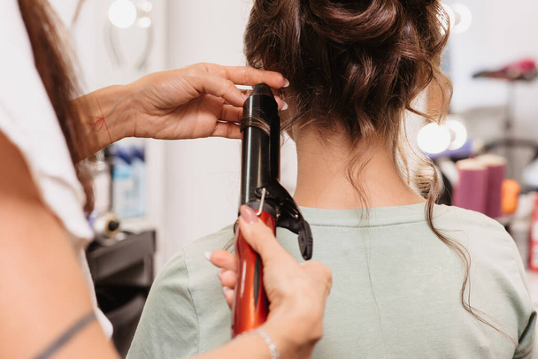 Shooting in a beauty salon. A hair stylist makes a hairstyle for a young dark-haired girl with a curling iron - Photo, Image