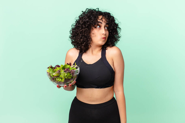 pretty arab woman shrugging, feeling confused and uncertain and holding a salad. diet concept - Foto, Imagen