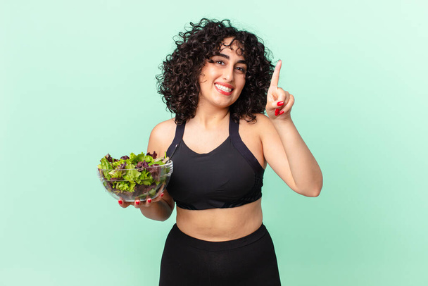 pretty arab woman smiling and looking friendly, showing number one and holding a salad. diet concept - Foto, afbeelding