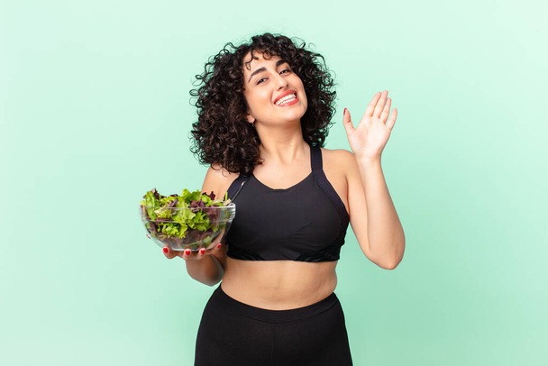 pretty arab woman smiling happily, waving hand, welcoming and greeting you and holding a salad. diet concept - Fotó, kép