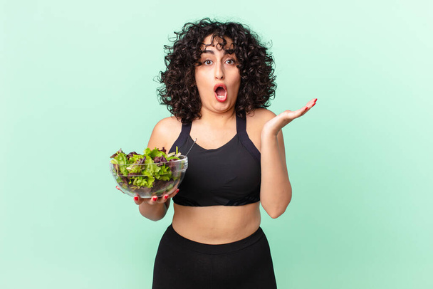 pretty arab woman looking surprised and shocked, with jaw dropped holding an object and holding a salad. diet concept - Zdjęcie, obraz