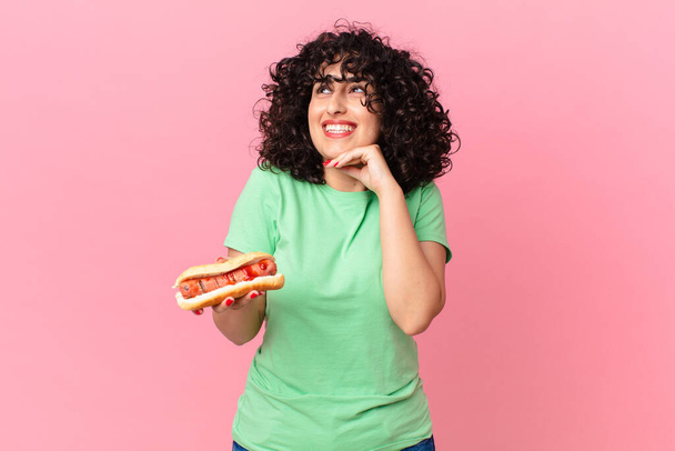 pretty arab woman smiling happily and daydreaming or doubting and holding a hot dog - Foto, Imagem