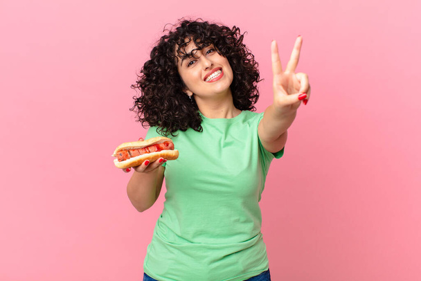 pretty arab woman smiling and looking happy, gesturing victory or peace and holding a hot dog - Foto, Imagem