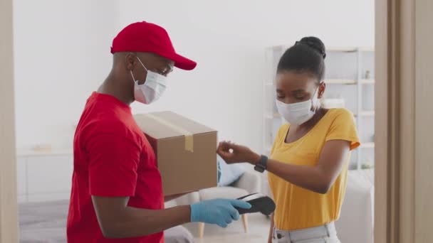 Młoda Afroamerykanka płaci za dostawę bezdotykową płatnością i przyjmowaniem zamówienia, obie w maskach ochronnych - Materiał filmowy, wideo