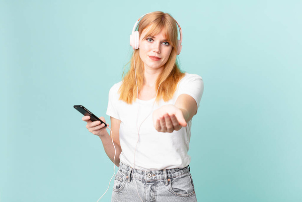 pretty red head woman smiling happily with friendly and  offering and showing a concept and listening music with headphones - Photo, Image