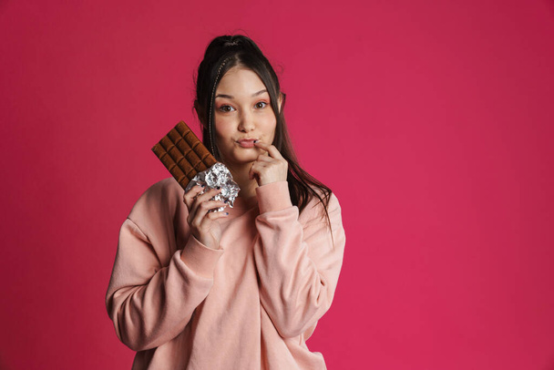 Pensive tempted young woman holding chocolate bar standing over pink background, thinking - Фото, изображение