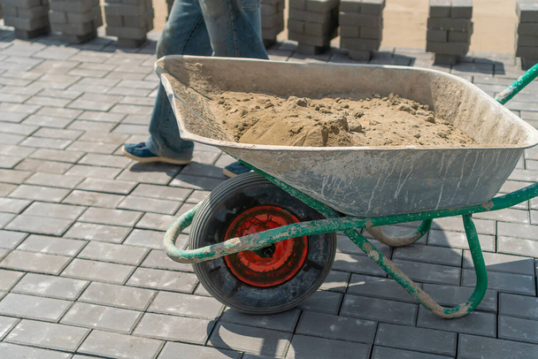 Detailní záběr dělníka s lopatou a plněním kolečka pískem na stavbu chodníku. Lopata a vozík na kolech s pískem na výstavbě silnic, chodníků - Fotografie, Obrázek