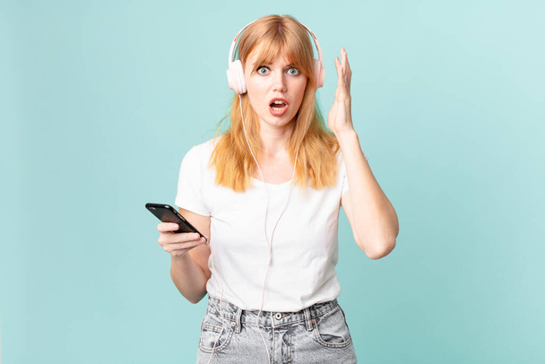 pretty red head woman screaming with hands up in the air and listening music with headphones - Foto, Imagen