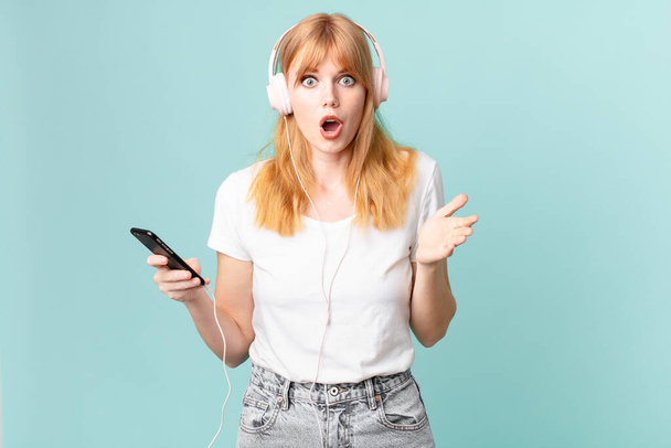 pretty red head woman feeling extremely shocked and surprised and listening music with headphones - Fotó, kép