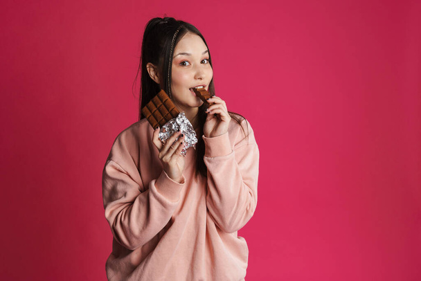 Happy young woman biting chocolate bar over pink wall background - Photo, image