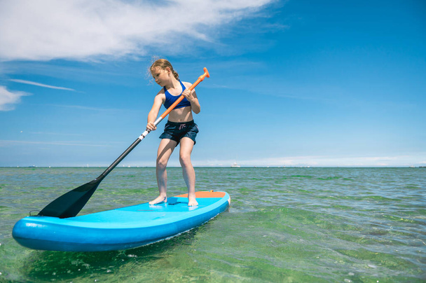 Cute little girl ecercise paddling on SUP board in sea on beautiful sunny summer day while summer holidays - Foto, imagen