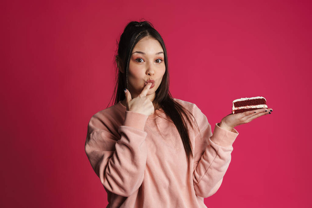 Asian happy woman licking her finger while posing with cake isolated over purple wall - Fotó, kép