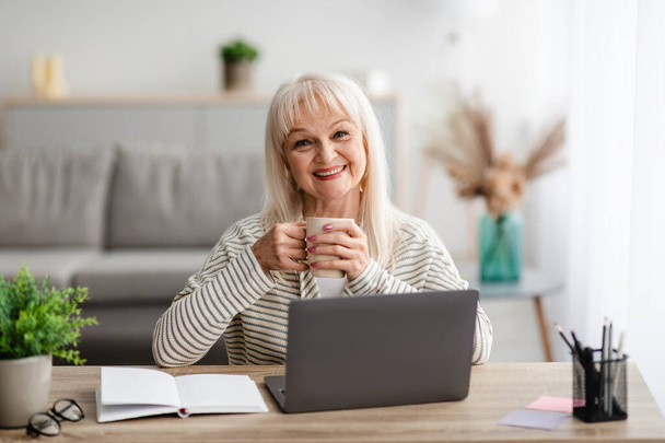 Vanhempi nainen istuu pöydän ääressä juomassa kahvia tietokoneella - Valokuva, kuva