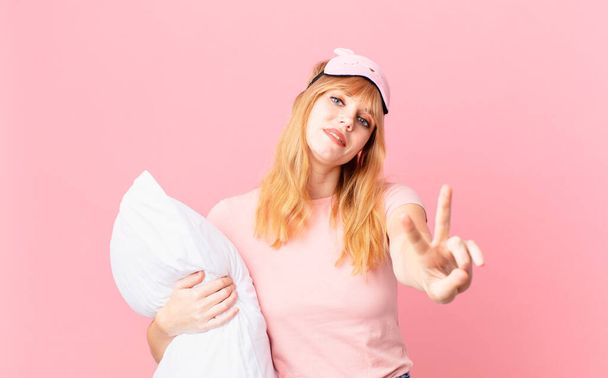 pretty red head woman smiling and looking happy, gesturing victory or peace. wearing pajamas and holding a pillow - Photo, image
