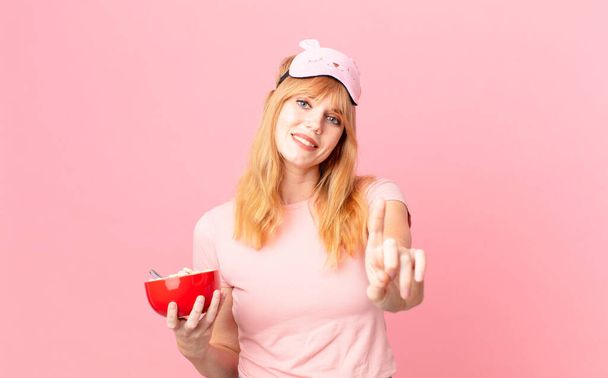 pretty red head woman smiling proudly and confidently making number one wearing pajamas and holding a flakes bowl - Фото, зображення