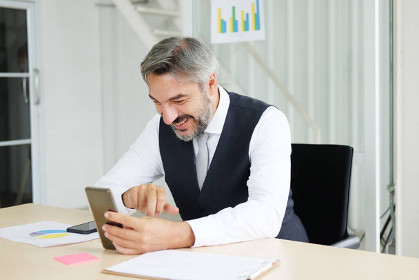 Χαμογελώντας έξυπνος Καυκάσιος ανώτερος διευθυντής και επιχειρηματίας με μαύρο κοστούμι χαλαρώνοντας στα μέσα κοινωνικής δικτύωσης online και μιλώντας, καλώντας με επιχειρηματίες στο smartphone στο χώρο εργασίας στο γραφείο - Φωτογραφία, εικόνα