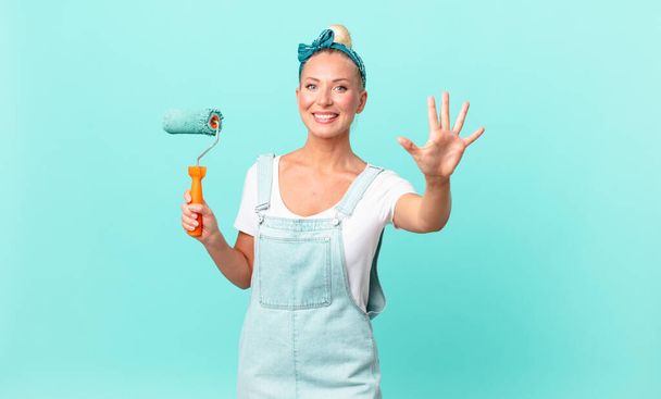 pretty blond woman smiling and looking friendly, showing number five and painting a wall - Foto, Bild