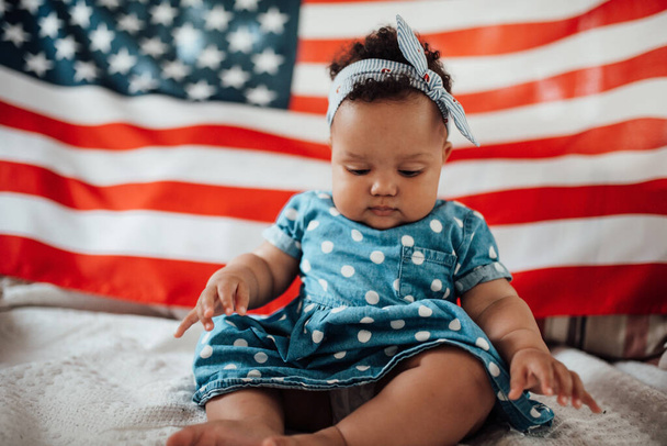 Lovely little girl in blue dress sitting on sofa with American flag background   - Фото, изображение