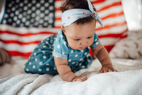 Mooi klein meisje in blauwe jurk zitten op de bank met Amerikaanse vlag achtergrond   - Foto, afbeelding