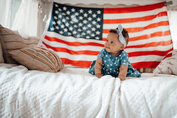Lovely little girl in blue dress sitting on sofa with American flag background   - Photo, Image