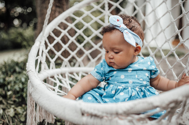 Outdoor portret cute little girl w niebieskiej sukience siedzi na krześle wiklinowym - Zdjęcie, obraz