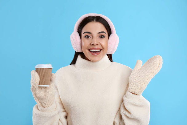 Beautiful young woman in earmuffs with cup of drink on light blue background - Fotografie, Obrázek
