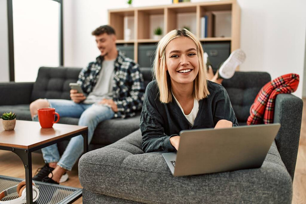 若い白人のカップルは、自宅でソファに座ってノートパソコンやスマートフォンを使用して幸せな笑顔. - 写真・画像