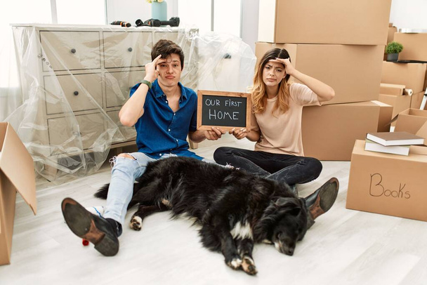 Junges kaukasisches Paar mit Hund, das unsere erste Haustafel im neuen Haus hält, besorgt und gestresst über ein Problem mit der Hand auf der Stirn, nervös und krisenängstlich  - Foto, Bild