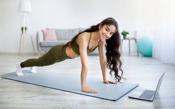 Smiling young Indian lady doing plank, strengthening her arms and body in front of laptop at home - Zdjęcie, obraz