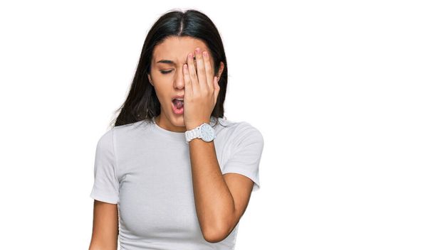 Jong Latijns-Amerikaans meisje draagt casual wit t-shirt geeuwen moe bedekken half gezicht, oog en mond met de hand. gezicht doet pijn in pijn.  - Foto, afbeelding