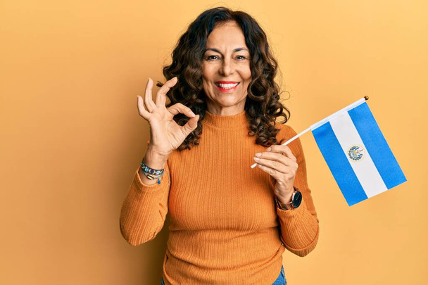 Mulher hispânica de meia idade segurando a bandeira de El Salvador fazendo sinal ok com os dedos, sorrindo simpática gesto excelente símbolo  - Foto, Imagem