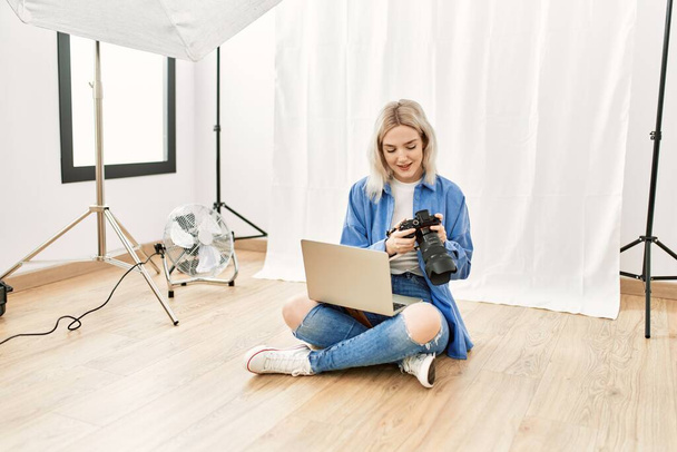 Beautiful blonde woman working as professional photographer at photography studio sitting on the floor checking photos on computer laptop - Photo, Image