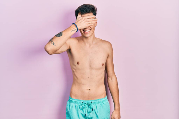 Jeune homme hispanique portant des maillots de bain torse nu souriant et riant avec la main sur le visage couvrant les yeux pour la surprise. concept aveugle.  - Photo, image