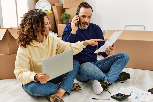 Middle age hispanic couple controlling family economy using laptop. Sitting on the floor talking on the smartphone at new home. - Photo, Image