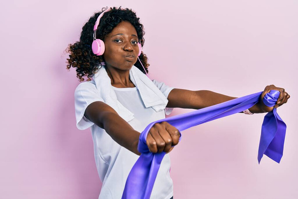 Young african american woman training arm resistance with elastic arm bands using headphones puffing cheeks with funny face. mouth inflated with air, catching air.  - Foto, immagini