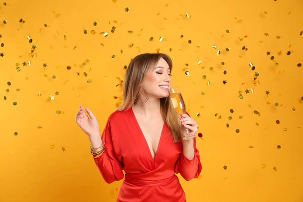 Happy young woman with glass of champagne and confetti on yellow background - Fotó, kép